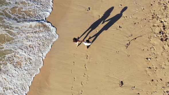 Couple in love holding hands walking on beach washed by sea flows, making long shadows by morning su