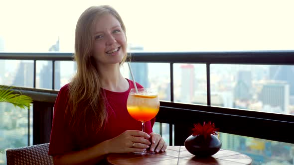 Young Woman with Cocktail Drink at High Rooftop Luxury Restaurant Looking at Camera Smiling