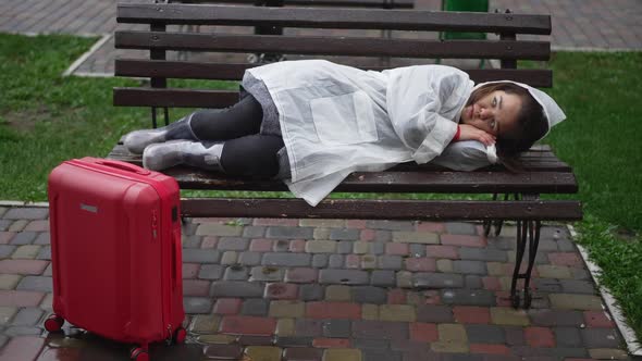 Wide Shot Sad Hopeless Little Woman in Rain Coat Lying on Bench Outdoors with Travel Bag Standing