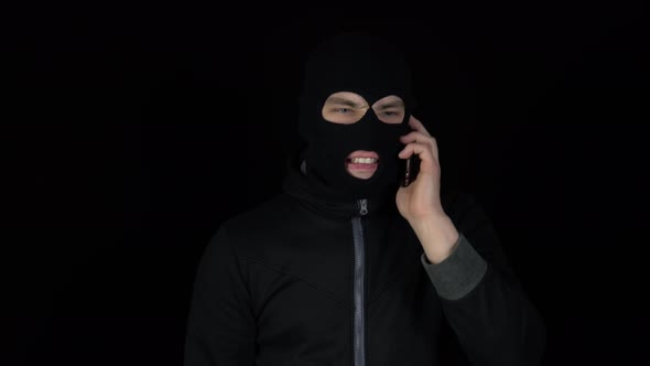 A Man in a Balaclava Mask Is Talking on a Phone. A Thug Screaming on the Phone on a Black Background