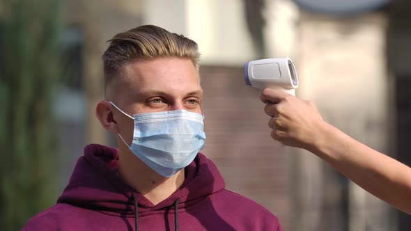 Portrait of Young Man in Medical Mask Whose Temperature Is Measured with a Non Contact Thermometer