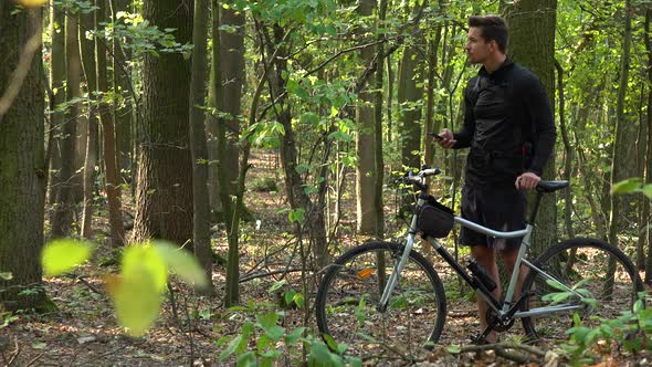 A Cyclist Stands with a Bike in a Forest and Works on a Smartphone