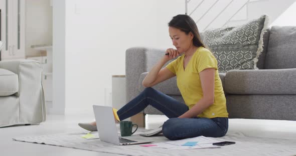 Focused asian woman sitting on floor and working remotely from home with smartphone and laptop