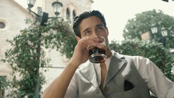 Attractive young man in a suit on a summer terrace drinking soda and waiting for someone