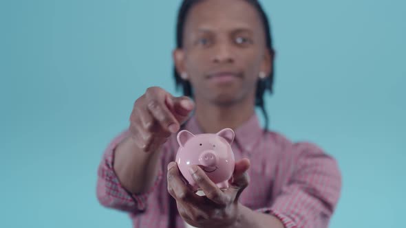 Black man with dreadlocks holds a piggy bank in hand and puts the coins close-up on a blue backgroun