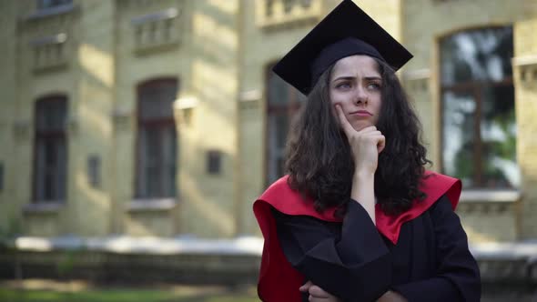 Thoughtful Young Beautiful Woman in Mortarboard Cap and Graduation Toga Standing at University