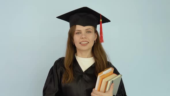 A Young Caucasian Woman in a Black Robe and a Master's Hat Stands Straight Holding Textbooks with