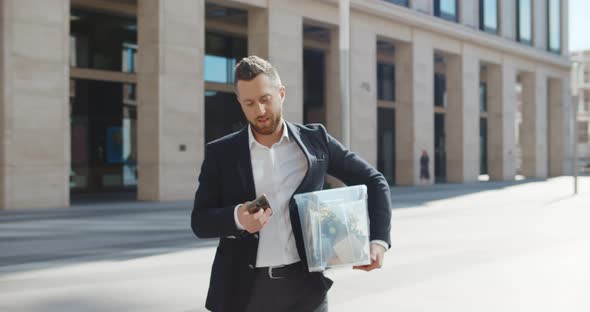 Upset Businessman in Suit Walking Outdoors with Box of Stuff Using Smartphone