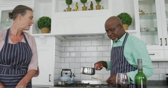 Smiling senior diverse couple wearing blue aprons and cooking in kitchen