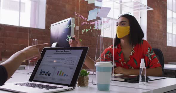 Mixed race businesswoman writing on sneez screen sitting in front of computer wearing face mask