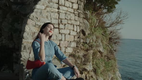 Young smiling brunette woman with red lipstick in denim overalls  sits on sunlights