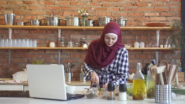 Muslim Woman in a Hijab in the Kitchen Looking at a Laptop Training Video How To Cook a Dish