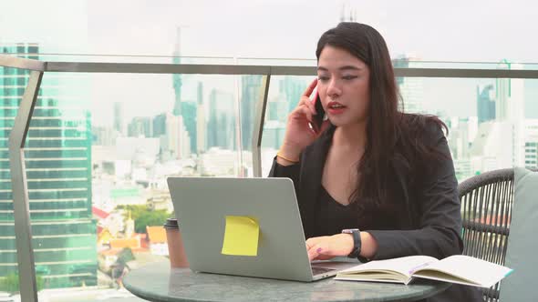 Asian woman talking by phone during working with laptop  along with writing in a notebook in cafe