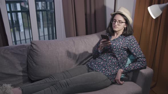 Girl in Glasses and a Hat Reads Messages in Mobile Phone While Sitting on the Couch in the Evening