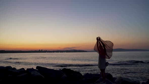 Girl Stands on Beach and Holds Scarf Over Her Head Against Sunset Over Sea Horizon