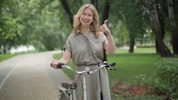 Portrait of Joyful Blond Middle Aged Woman Standing with Bike Looking at Camera Smiling and Showing