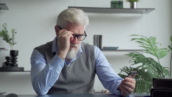 An Elderly Man with a Gray Beard Uses a Smartphone in the Office