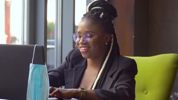 Young Black Woman Use Laptop and Looking at the Camera. Medical Disposable Mask Hanging on the