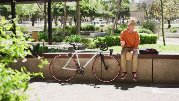 Thoughtful albino african american man with dreadlocks sitting in park with bike using smartphone