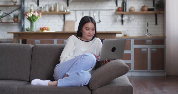 Asian Freelancer in White Shirt Lying on Couch with Laptop Typing on Keyboard and Blinking While