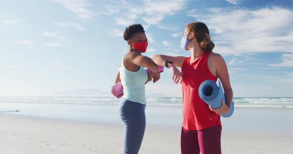 Two diverse female friends wearing face masks holding yoga mats at the beach touching elbows