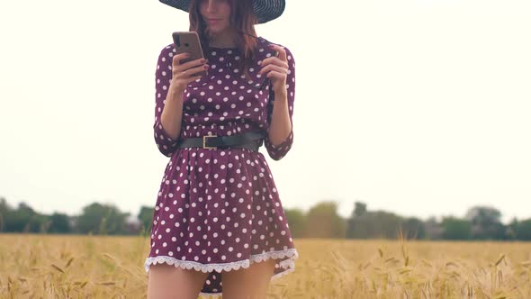 Beautiful Young Hippie Woman in the Wheat Field at Sunset Uses a Smartphone