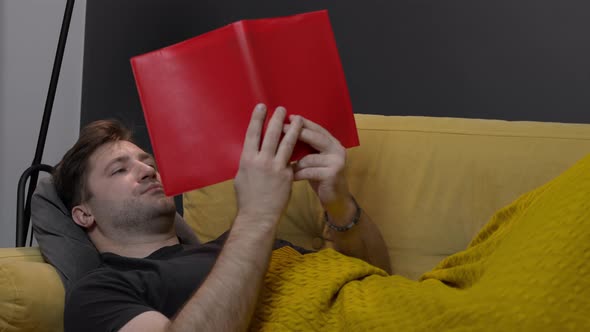 Young Unshaven Handsome Guy Reads Book Liyng on Comfortable Yellow Sofa