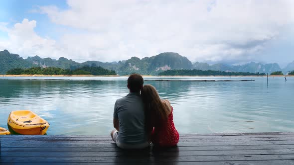 Happy Romantic Couple Sitting Together on Wooden Bridge on Green Mountains Lake