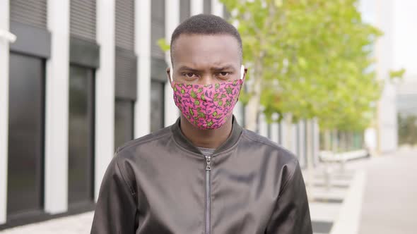 A Young Black Man in a Face Mask Looks at the Camera - an Office Building in the Blurry Background