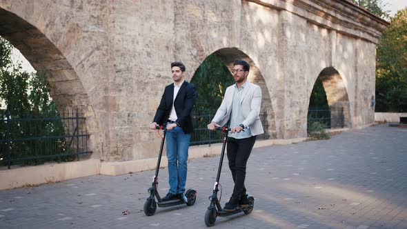 Two Handsome Young Businessman Riding an Electric Scooters on the Road in City Park Slow Motion