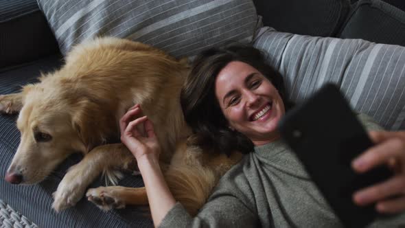 Smiling caucasian woman taking selfie with smartphone, stroking her pet dog on sofa next to her