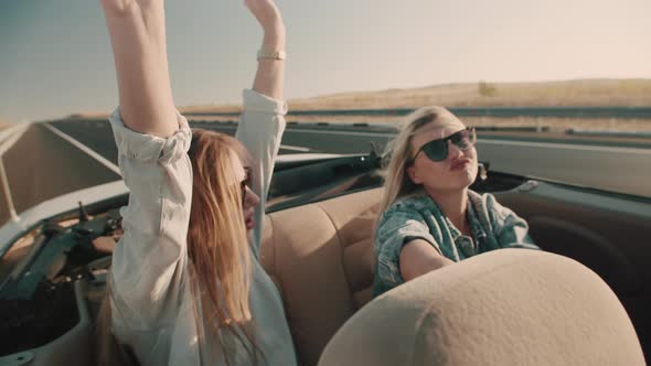 Two young women in sunglasses sing and dance with their hands sitting in back seat of convertible.