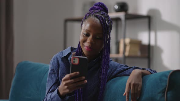Cheerful Afroamerican Young Woman is Viewing Funny Video or Movie on Screen of Smartphone Sitting