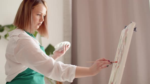 Woman is Working Intently in Workshop Painting on Canvas with Brush and Holding Palette with Paints