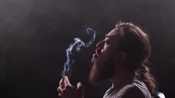 A Guy with a Long Beard and Oblique Smokes a Cigarette on a Black Background