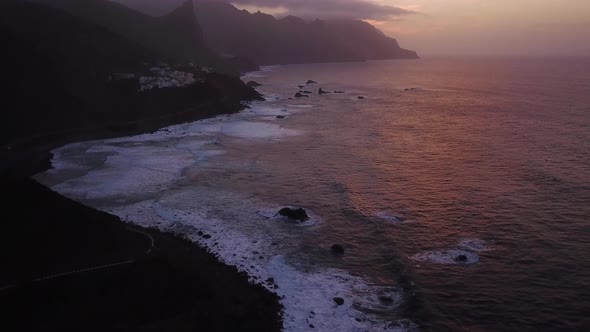 Aerial revealing shot of the volcanic cliffs of Tenerife with waves splashing during the sunset.