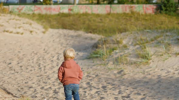 Blond Boy in a Hoodie Walks on the Sand