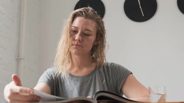 Close Up Footage Beautiful Blonde in Gray T-shirt Sits at Table and Reads Magazine. Drinks Water