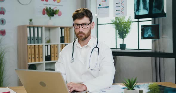 Doctor in Glasses Working on Laptop and then Looking into Camera with Sincerely Smile