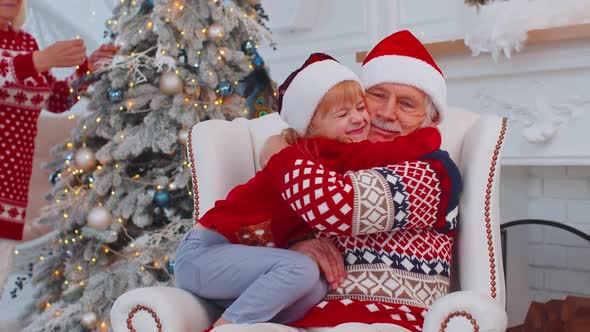 Granddaughter Child Fixes Christmas Santa Claus Hat on Happy Senior Old Smiling Grandfather at Home