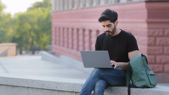 Millennial Student Using Laptop Typing on Keyboard Outdoors Looks at Monitor Scared Raises His Hand