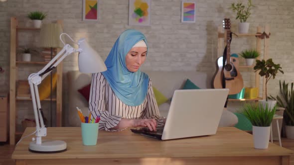 Young Muslim Woman in a Hijab with a Scar From a Burn on Her Face Works at a Laptop