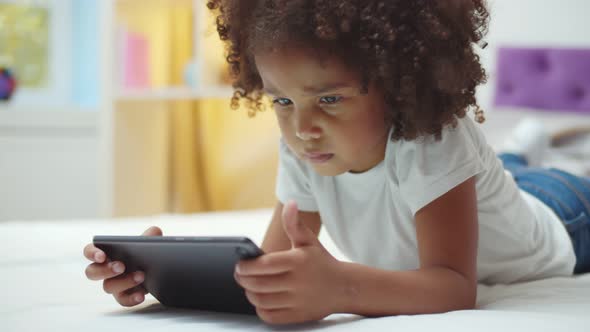 Adorable Afro Kid Girl Using Digital Tablet Watching Cartoons Lying on Bed at Home