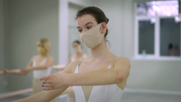 Portrait of Serious Ballerina in Covid Face Mask Rehearsing Hand Position with People at Barre in