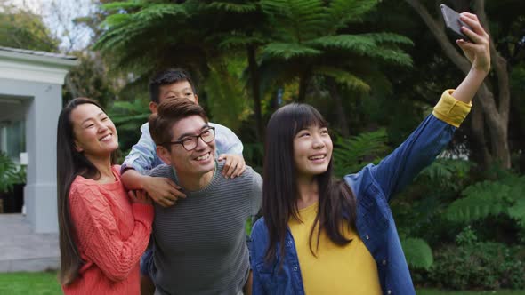 Happy asian daughter taking selfie with happy mother, and father piggybacking brother, in garden