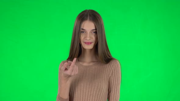 Portrait of Slender Girl with Long Hair Is Waving Hand and Showing Gesture Come Here on a Green