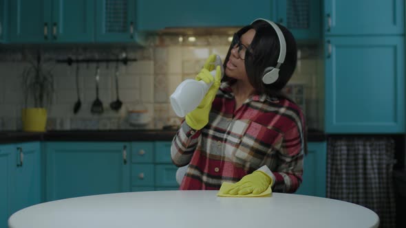 Young Adult African American Woman Listening Music in Headphones While Cleaning Table with Cleaning