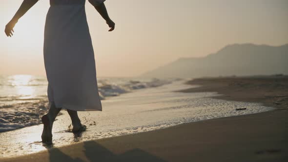 Happy tourist in white dress walking by the sea on the beach against the backdrop of sunset