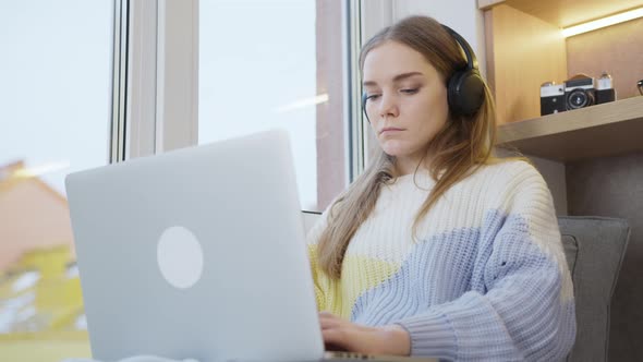 Young woman sitting by the window at home in headphones, working on a laptop. It's snowing outside.