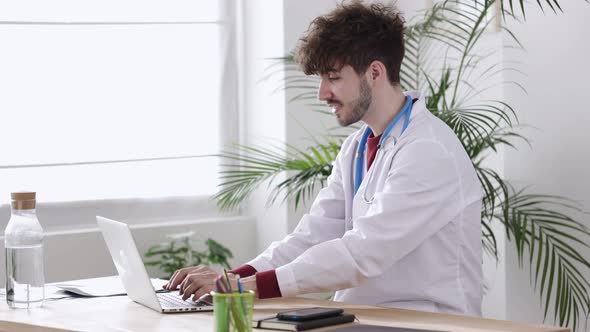 Young Man Doctor Working on Laptop at Desk in Medical Office